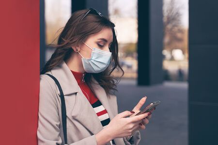 Young woman wearing protection face mask against coronavirus COVID-19 aka 2019-nCoV is looking for information about the outbreak on hwe smartphone while standing on the street. Concept of coronavirus quarantine and pandemic.の写真素材
