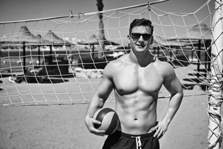 Handsome fit strong athletic young man in swimming trunks holds a volleyball ball on a beach sand volleyball court with net in a sea resort. Fitness model or sportsman on vacation. Black and white.の写真素材