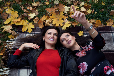Two beautiful adult sisters are laying down on a plaid carpet in autumn park with yellow and orange...