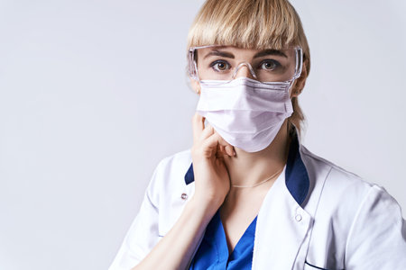 Confident blonde female doctor wearing protective face mask. Portrait on light grey background.