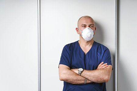 Experienced male massage therapist masseur in his medical office standing by a massage table wearing professional uniform and disposable standard N95 filtering facepiece respirator face mask.の写真素材