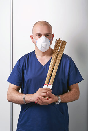 Experienced male massage therapist masseur in his medical office wearing professional uniform and disposable respirator face mask. Holds traditional bamboo massage sticks.の写真素材