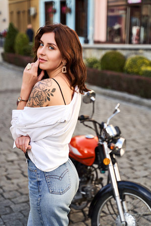 Young beautiful woman shows her bare shoulder with a tattoo standing by a bike on the streetの写真素材