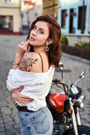 Young beautiful woman shows her bare shoulder with a tattoo standing by a bike on the streetの写真素材