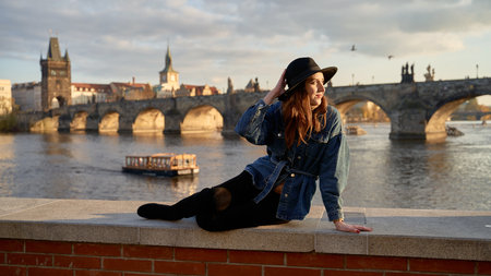 Stylish beautiful young woman wearing black hat sitting on Vltava river shore in Prague with Charles Bridge on background. Elegant retro lady fine art portrait.の写真素材