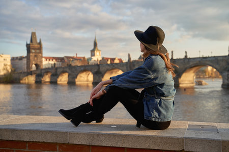 Stylish beautiful young woman wearing black hat sitting on Vltava river shore in Prague with Charles Bridge on background. Elegant retro lady fine art portrait.の写真素材
