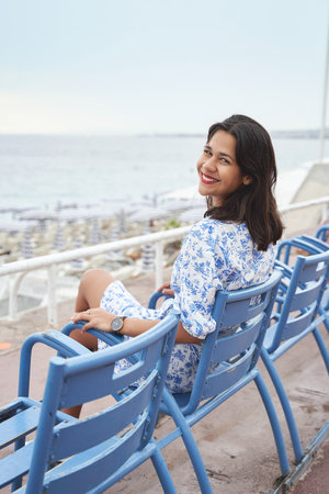 Beautiful smiling young mixed race woman sitting at one of the famous blue chairs at the Promenade des Anglais in Nice, Franceの写真素材