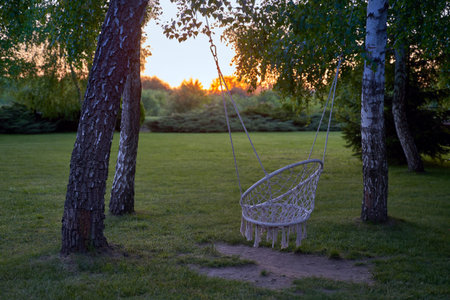 Sunset garden. Bohemian style hammock hanging swing chair with tassels on ropes in a evening garden at duskの写真素材