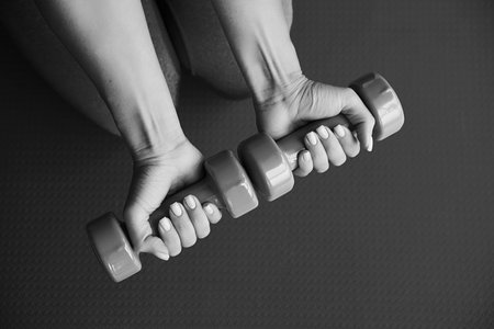 Black and white woman hands holding dumbbells on green sport mat background. Home exercise during pandemic. Workout in gym. Fitness concept.の写真素材