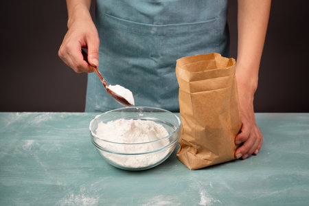 Baker puts wheat flour with a spoon in a bowl, paper bag with baking ingredient for bread, pizza or pastryの写真素材