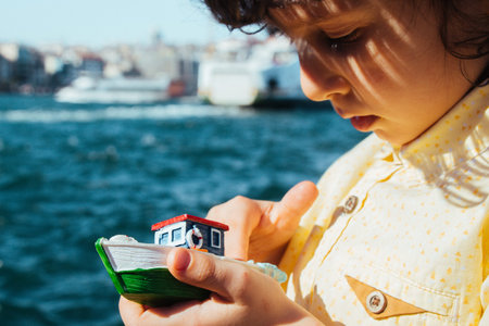 Child holding a boat model cheerfully, sea in thebackground.
