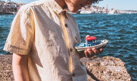 Child holding a boat model cheerfully, sea in thebackground.の写真素材