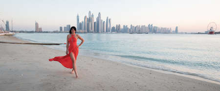 Beautiful woman with a red dress is walking on the beach in Dubai. In the background there is the skyline from Dubai Marinaの写真素材