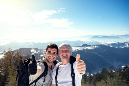 Cheerful young and middle aged man keeping thumb up on the top of the mountain. Direct sunlight, warmer highlights, shallow depth of field.の写真素材