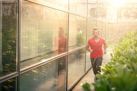 Young male jogging next to modern sport center, listening music with his headphones on. Wearing shirt and tracksuits. Selective focus, toned image, direct sunlight, lens flare.の写真素材
