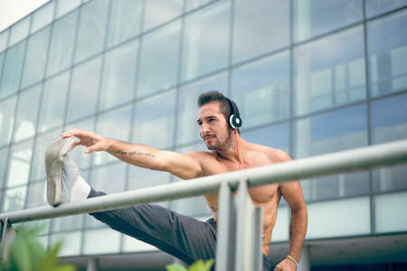 Young man listening to the music and stretching his leg after a heavy workout. Wearing tracksuits without shirt. Selective focus.の写真素材