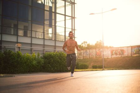 Young male jogging next to modern sport center, listening music with his headphones on. Wearing tracksuits without shirt. Toned image, direct sunlight, lens flare.の写真素材