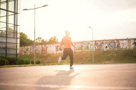 Young male jogging next to modern sport center, listening music with his headphones on. Wearing tracksuits without shirt. Toned image, direct sunlight, lens flare.の写真素材