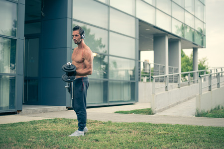 Young man listening to the music and working out with dumbbells in front of the modern sport centar. Wearing tracksuits without shirt. Selective focusの写真素材