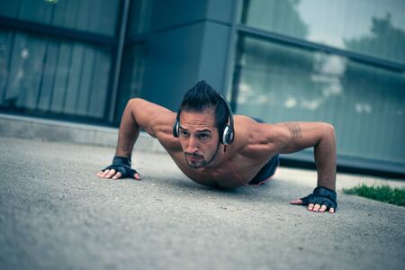 Young male listening to the music and doing push ups in front of the modern sport centar. Selective focus.の写真素材