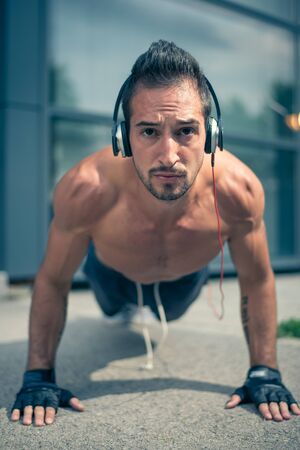 Young male listening to the music and doing push ups in front of the modern sport centar. Selective focus.の写真素材