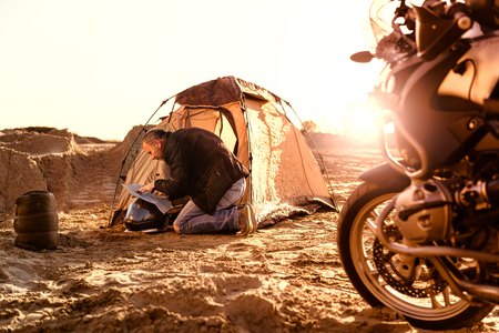 Middle aged motorcyclist relaxing after a long ride on motorbike. Checking his travel map in tent. Lens flare. Warm colors. Selective focus.の写真素材