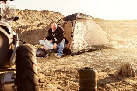 Middle aged motorcyclist relaxing after a long ride on motorbike. Checking his travel map in tent. Lens flare. Warm colors. Selective focus.の写真素材