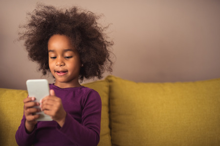 Curious little girl playing games on her mobile phone indoors.の写真素材