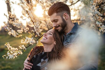 Photo of a couple enjoying in the park at sunset. The man is embracing his girlfriend from behind. They are both happy.の写真素材
