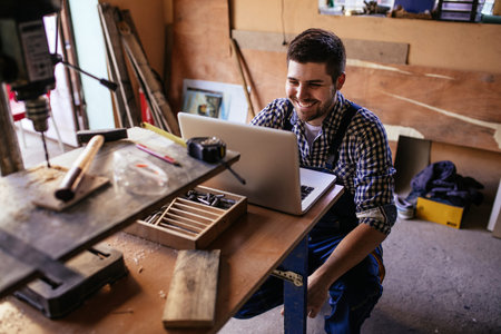 Portrait of a smiling young carpenter sitting at the table and using laptop in workshop.