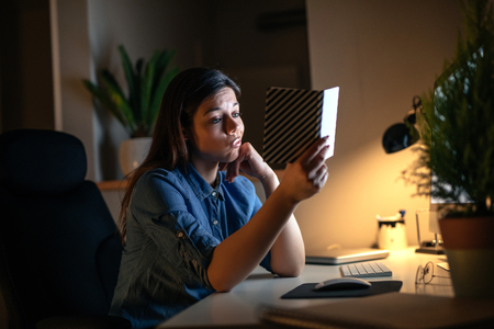 Portrait of a woman reading a notebook while sitting at the computer table in the evening.の写真素材