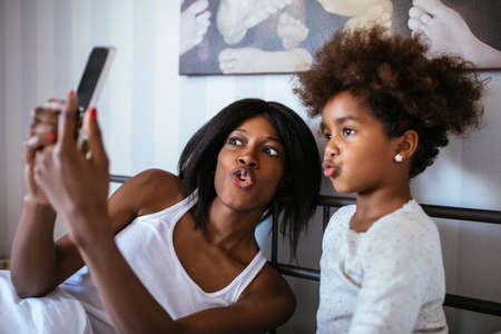Photo of african american mother and daughter taking a picture with mobile phone in the bed.の写真素材