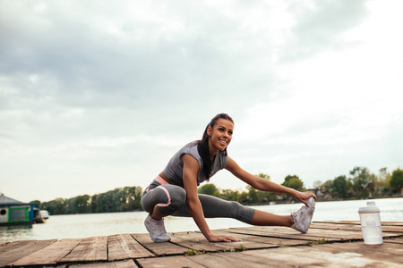 Full length portrait of an athlete young woman stretching outdoors, near the lake.の写真素材