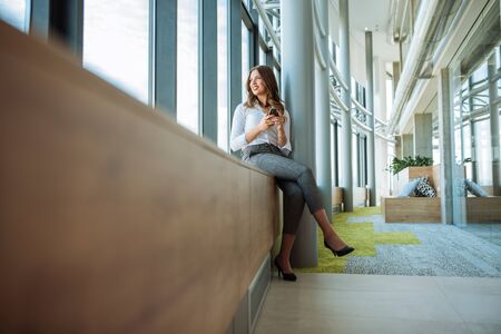 Full length portrait of a beautiful business woman sitting by the window and using mobile phone.の写真素材