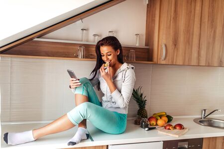 Shot of a healthy woman eating healthy food in the kitchen while using mobile phone.の写真素材