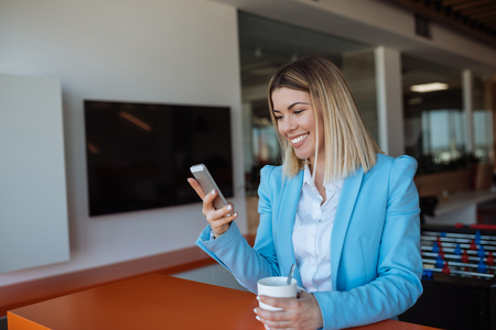 Shot of a smiling young business woman using mobile phone and drinking coffee.の写真素材