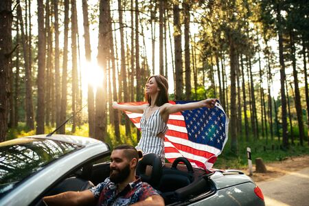Shot of a young couple enjoying riding in a car. Focus on the woman who is holding an american flag.の写真素材