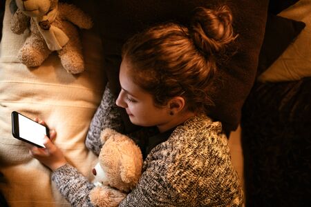 Shot from above of a young teenage girl using mobile phone while lying in bed and hugging a teddy bear.の写真素材