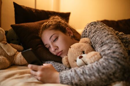 Photo of a sleepy teenage girl using mobile phone while lying in bed.の写真素材
