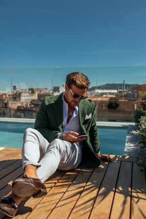 Handsome stylish man texting on the phone next to the pool.の写真素材