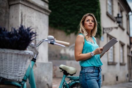 Photo of a young woman using digital tablet while standing next to her bicycle outdoors.の写真素材