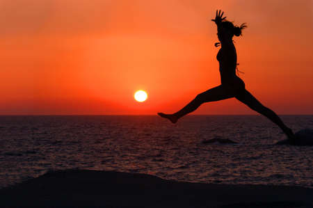 Young woman leaping towards the setting sunの写真素材