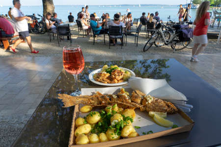 Balatonfured, Hungary, 06.10.2019 : Fish ad wine festival fried fish dishes and a glass of rose wine at lake balaton with people and lake background.のeditorial素材