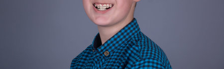 portrait of a teenager with plates on his teeth in a blue shirt on a gray studio background,...