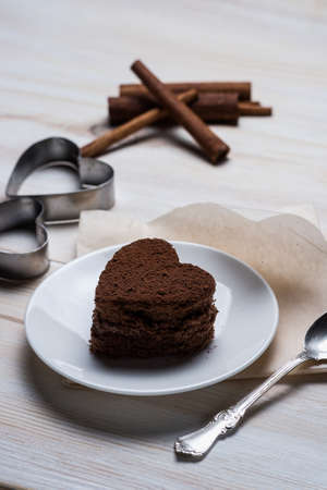 side view of a chocolate dessert biscuit in the shape of heart lying on a saucer next to a silver spoon and iron forms and cinnamon sticks on a white wooden background.の写真素材