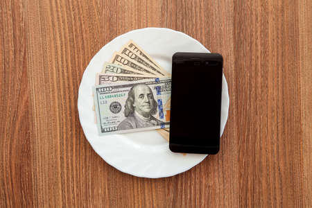 Wooden table on which there is a plate in which the denomination hundred, fifty, twenty, and ten dollars and a black business phone lies.の写真素材