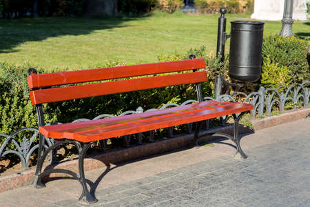 A bench with a garbage bin in a city park for relaxing on the background of green bushes.の写真素材