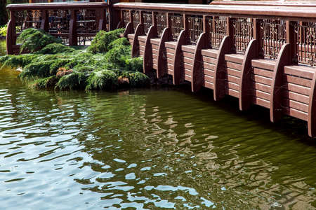 Water with waves and reflexes near a wooden bridge with railings and a green thuja bush.の写真素材