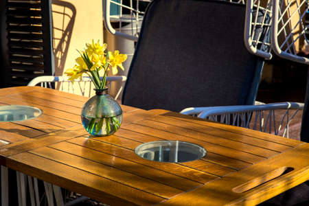 an empty street cafe with a wooden table made of brown boards with a glass insert; on the table is a ball-shaped vase with yellow flowers.の写真素材