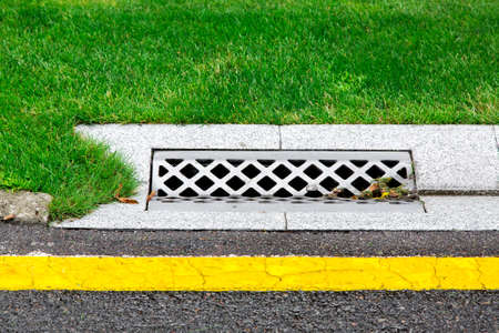 drainage system edge tray with concrete grate for rainwater drainage into the sewer on road with a yellow marking and a green lawn after rain.の写真素材
